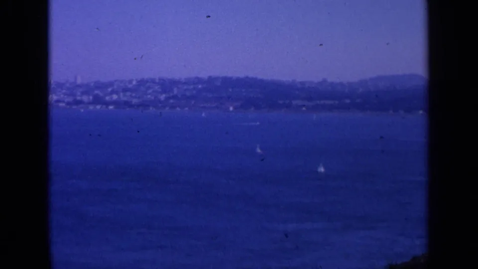 1963 San Francisco home-movie frame showing blue bay water, distant hills, and small sailboats through a dark 8mm film border.