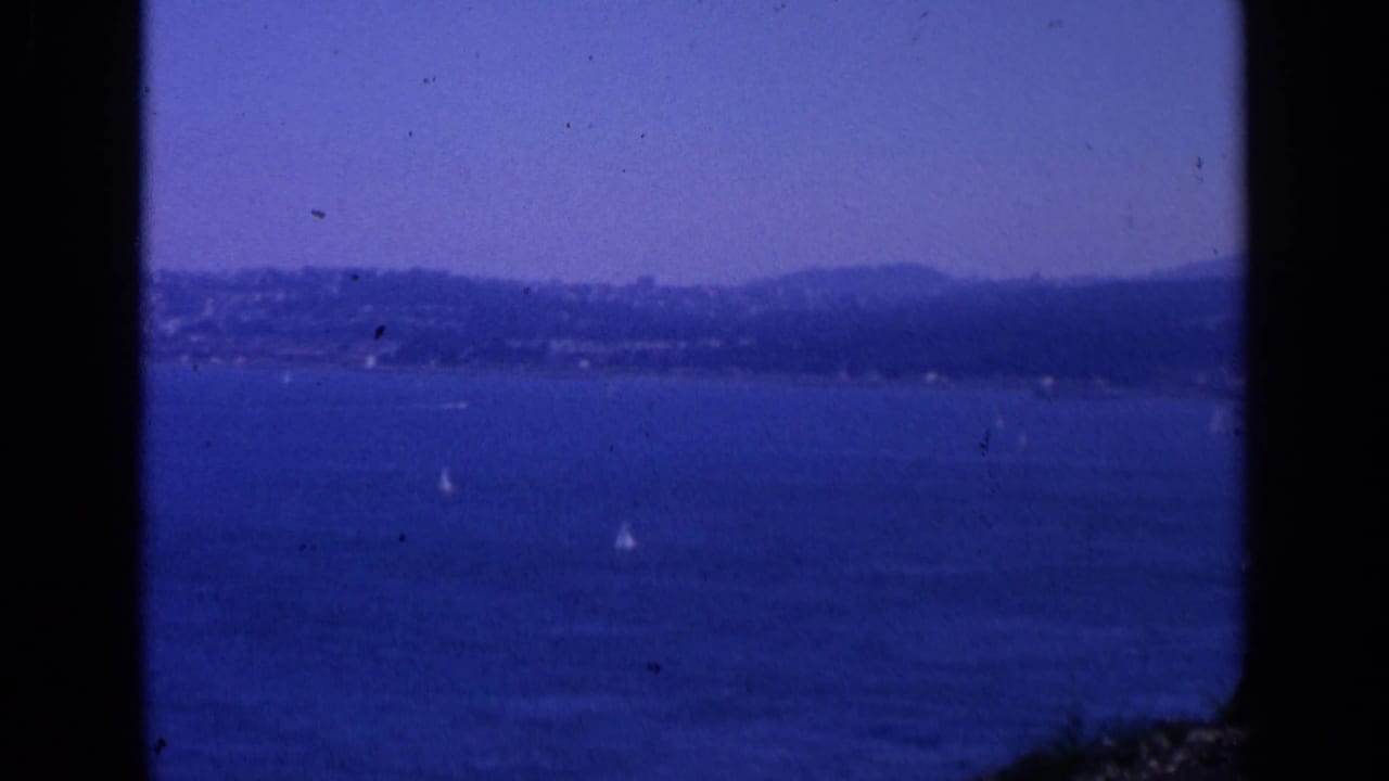 1963 San Francisco coastal view with hazy hills beyond blue water and scattered sailboats, framed by visible 8mm gate edges.
