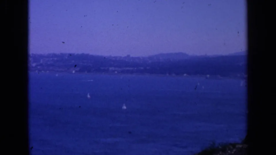 1963 San Francisco coastal view with hazy hills beyond blue water and scattered sailboats, framed by visible 8mm gate edges.