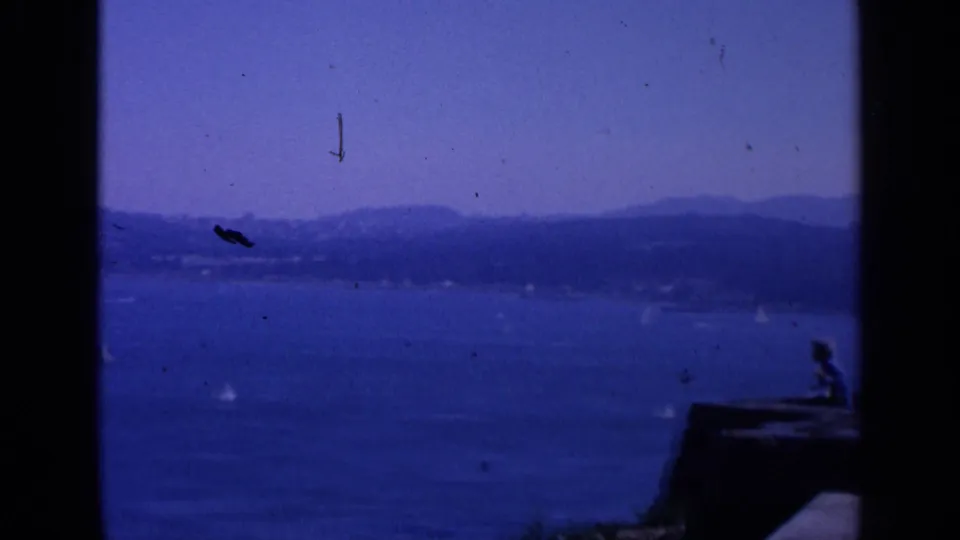 1963 San Francisco 8mm frame showing sailboats on blue water, hazy hills, and part of a rocky overlook entering the foreground.