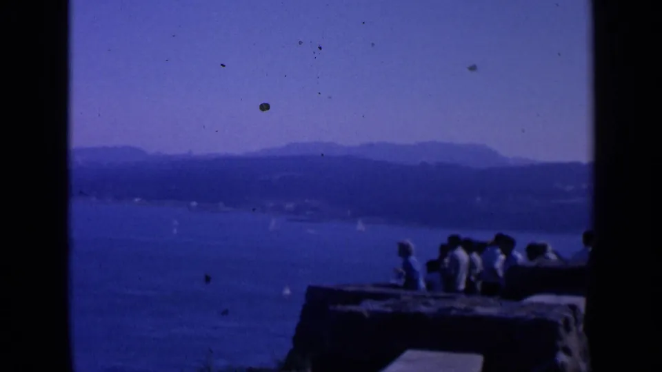 1963 San Francisco home-movie frame with a small group of visitors on a rocky overlook facing blue bay water, sailboats, and distant hills.