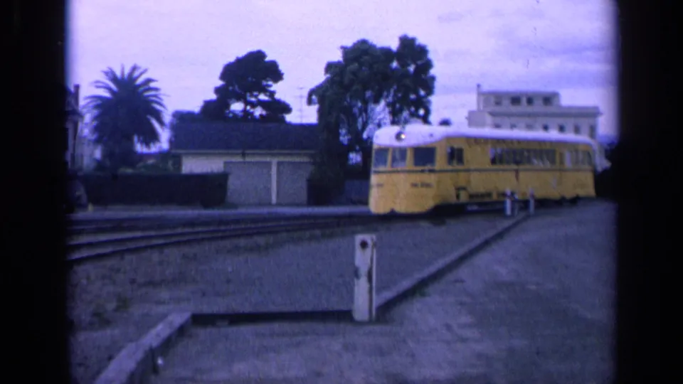 1963 San Francisco frame showing a yellow trolley moving on tracks beside low residential buildings and palm trees.