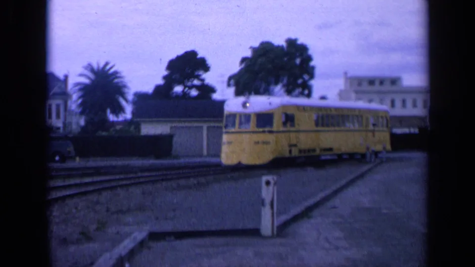 1963 San Francisco frame with a yellow trolley passing across rail tracks near houses, a parked car, palms, and a curb.