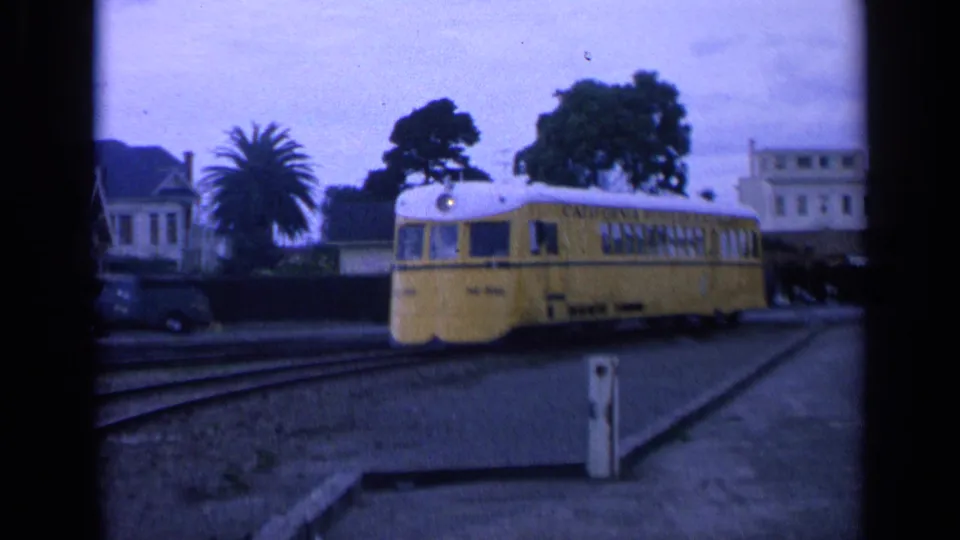 1963 San Francisco frame showing the yellow trolley moving past the crossing with residential buildings, palms, and trees behind it.
