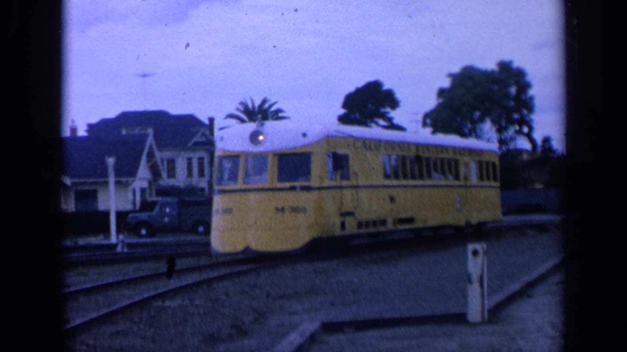 Yellow trolley car passing houses with tracks and trees in the foreground.