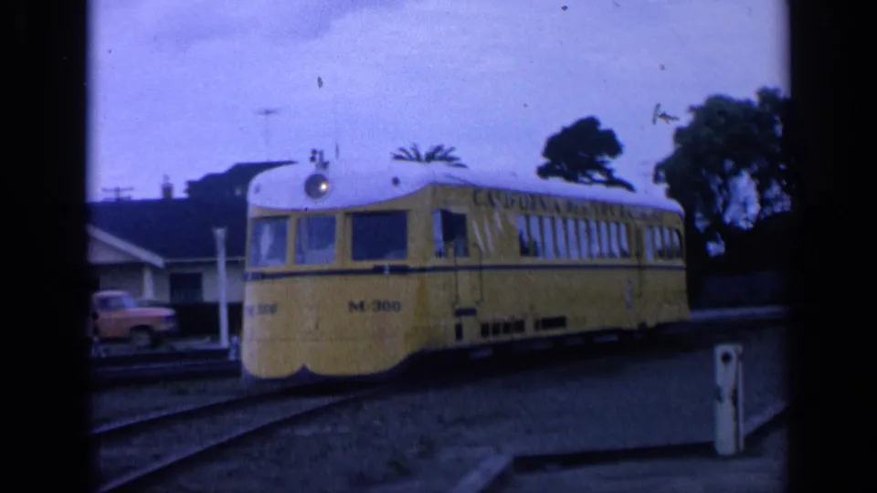 Yellow M-300 railcar in a 1963 San Francisco 8mm frame, with nearby houses, trees, and trackside details visible.
