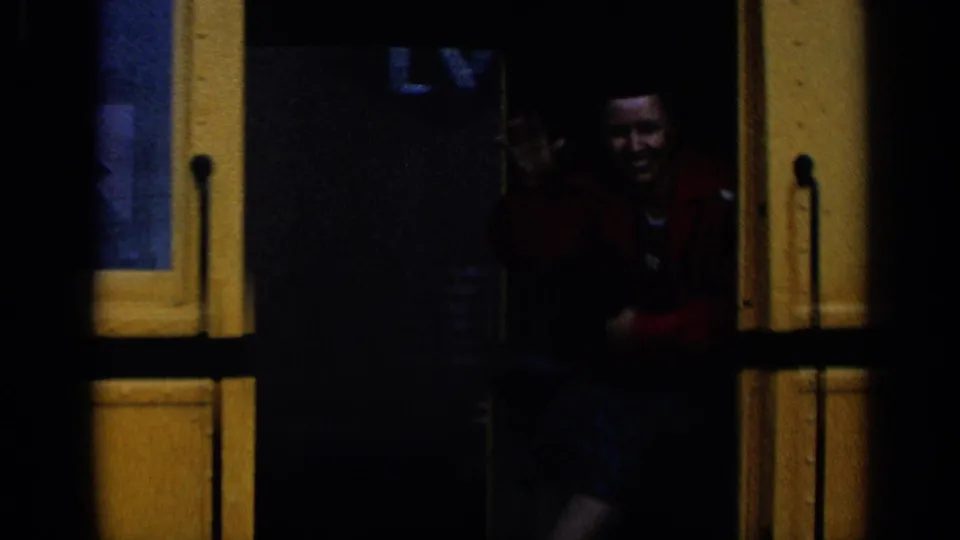 Passenger in a red jacket appears in the doorway of the yellow railcar, with facial features partly visible in shadow.