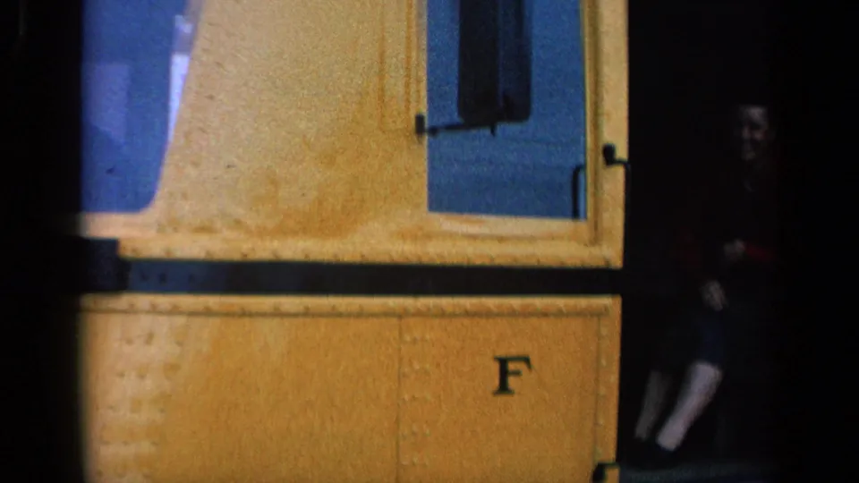 Close 8mm view of the yellow railcar side, showing windows, an open doorway, and a passenger partly seated in shadow.