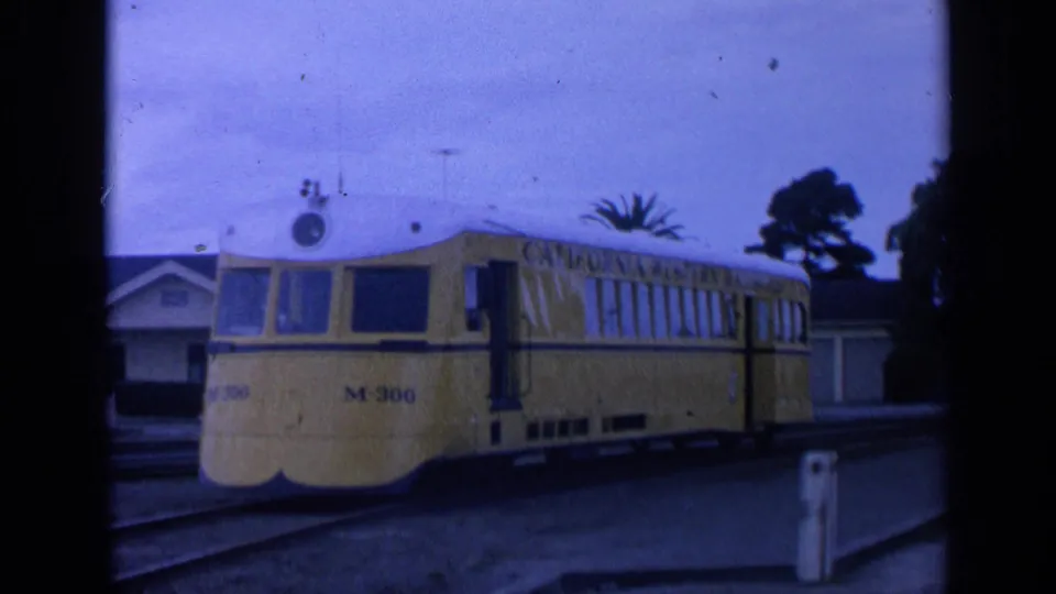 Yellow M-300 railcar at a California station, with side lettering, rounded front, and residential buildings in the background.