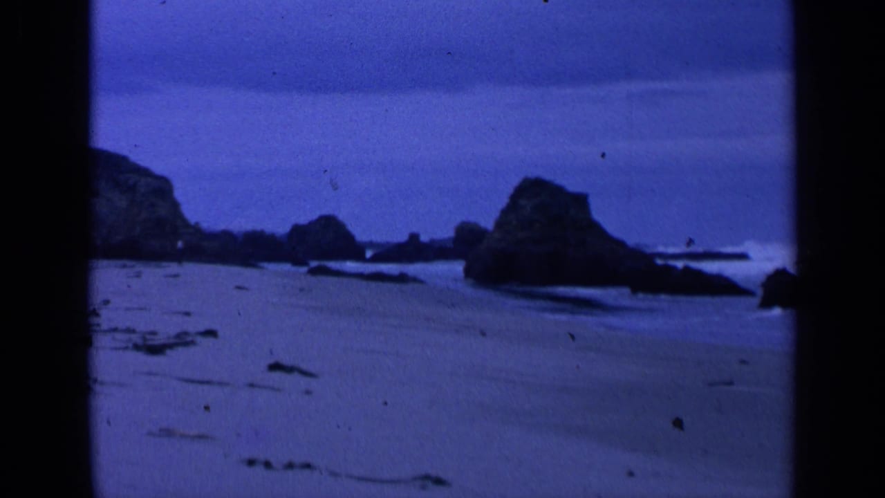 Blue-tinted beach view with wet sand, surf, and dark offshore rocks.