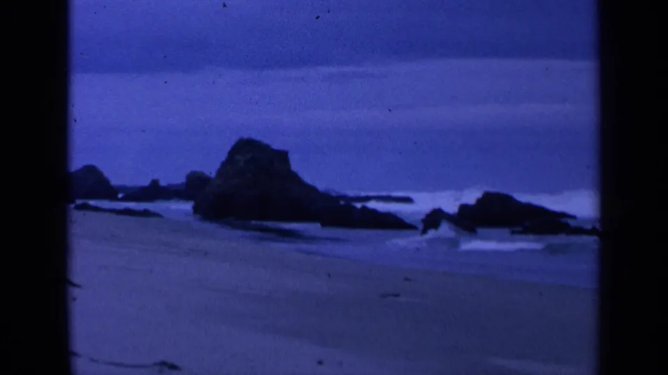 1963 San Francisco coastal frame with blue-tinted surf, sandy beach, and dark rock shapes along the waterline.