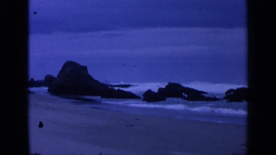 1963 San Francisco 8mm view of a large dark coastal rock, low surf, wet sand, and a muted blue sky.