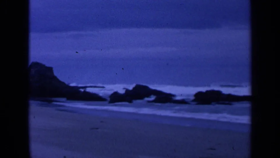 1963 San Francisco beach footage showing blue-toned surf washing across sand with dark rocks silhouetted offshore.