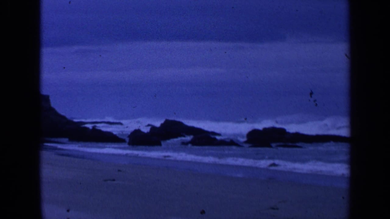 Blue-tinted surf breaks around dark boulders on a sandy beach.