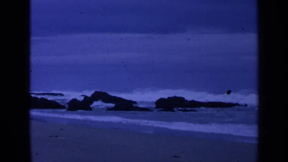 1963 San Francisco beach frame with low boulders, surf lines, wet sand, and blue monochrome film color.