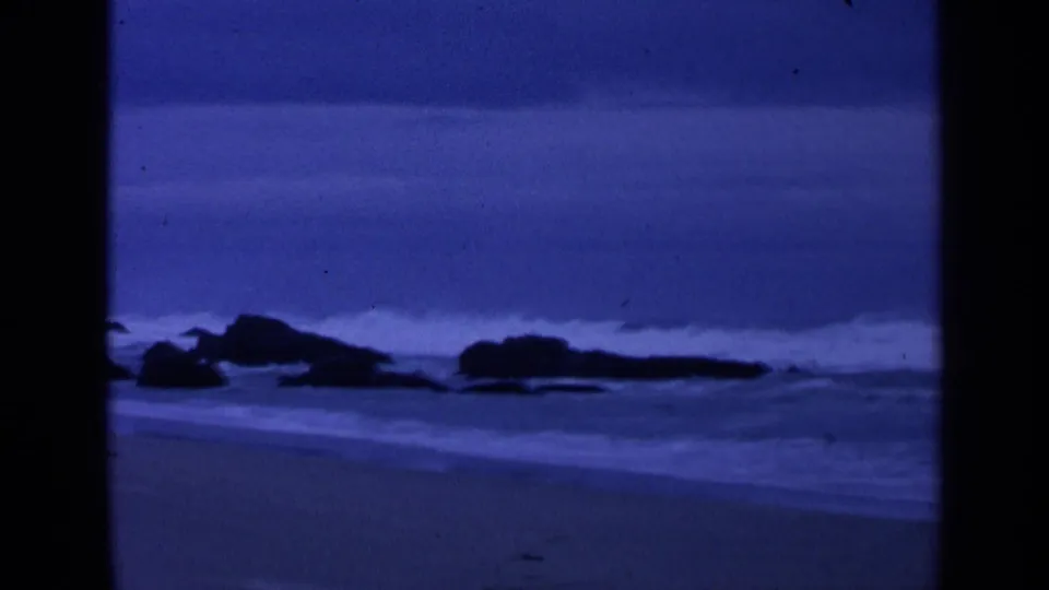 1963 San Francisco coastal frame showing dark boulders, white surf, wet beach sand, and heavy blue film tint.