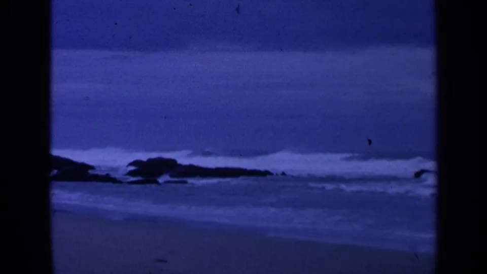 1963 San Francisco shoreline frame with blue-toned surf, dark rocks, wet sand, and visible 8mm frame edges.