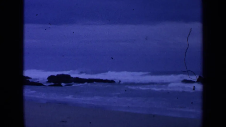 1963 8mm San Francisco-area coastal view showing blue surf, dark rocks, and a sandy shoreline in low light.