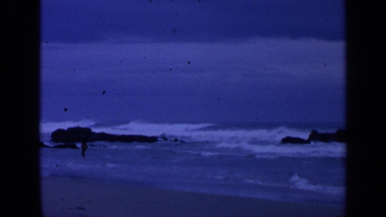 Surf rolls behind a dark rock formation on a blue shoreline.