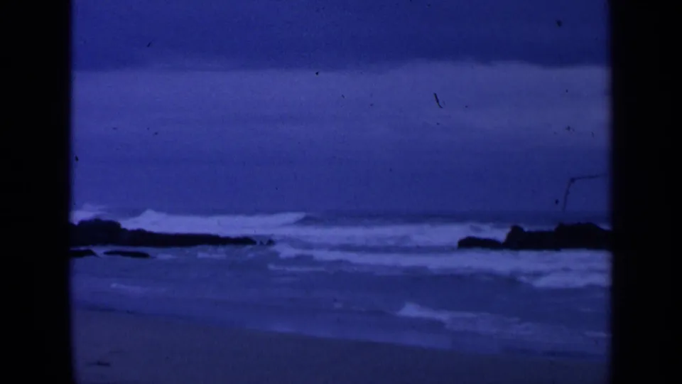 1963 8mm shoreline scene with white surf, dark rock silhouettes, and a blue-tinted sandy beach.