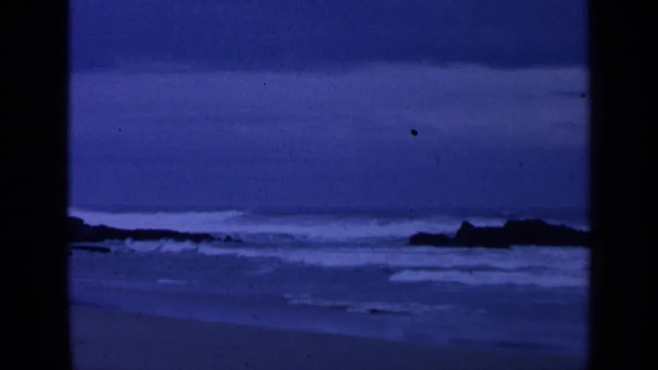 Blue low-light coastal frame from 1963 8mm footage, showing waves nearing sand and dark rocks at the frame edge.