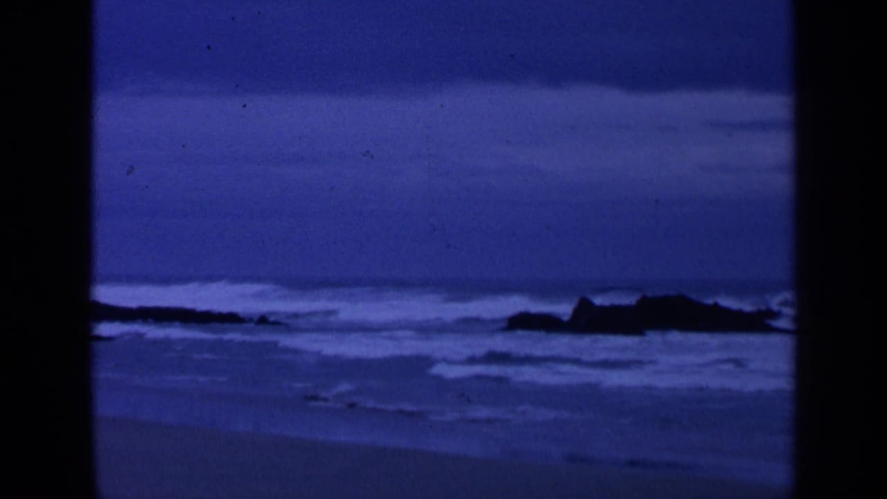 Blue-toned surf breaks around dark rocks near a sandy shore.