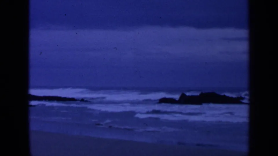 1963 San Francisco 8mm frame showing blue overcast surf breaking around dark rocks along the shoreline.