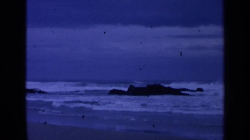 1963 San Francisco coastal frame with foamy waves, low rocks, and a cool blue overcast horizon.