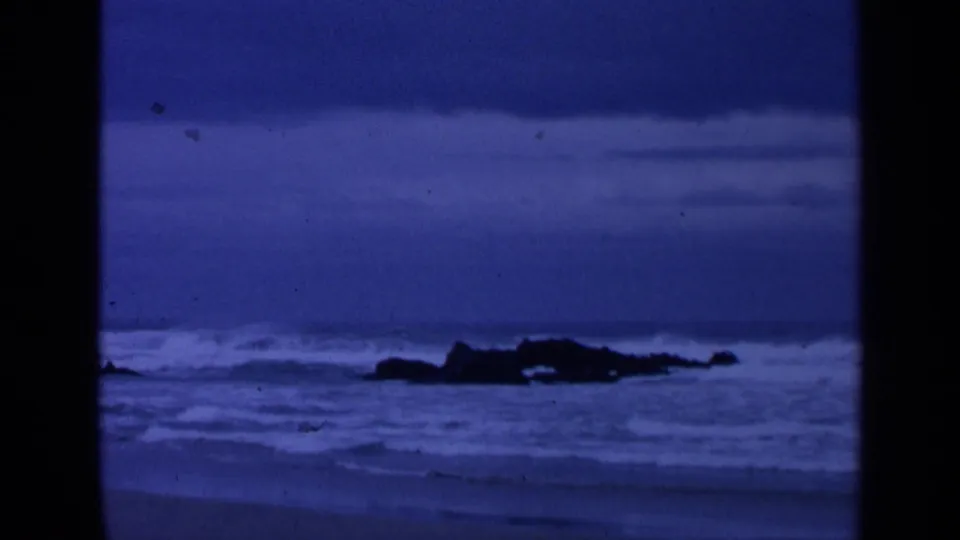 Blue-tinted 8mm view of San Francisco surf washing around dark shoreline rocks in 1963.