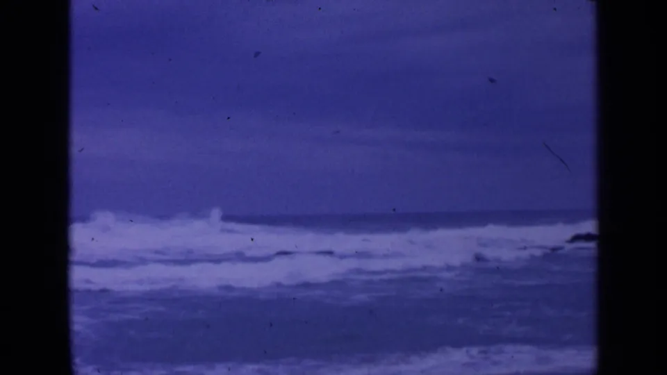 1963 coastal 8mm frame showing turbulent waves and foam bands, with heavy blue-purple film coloration.