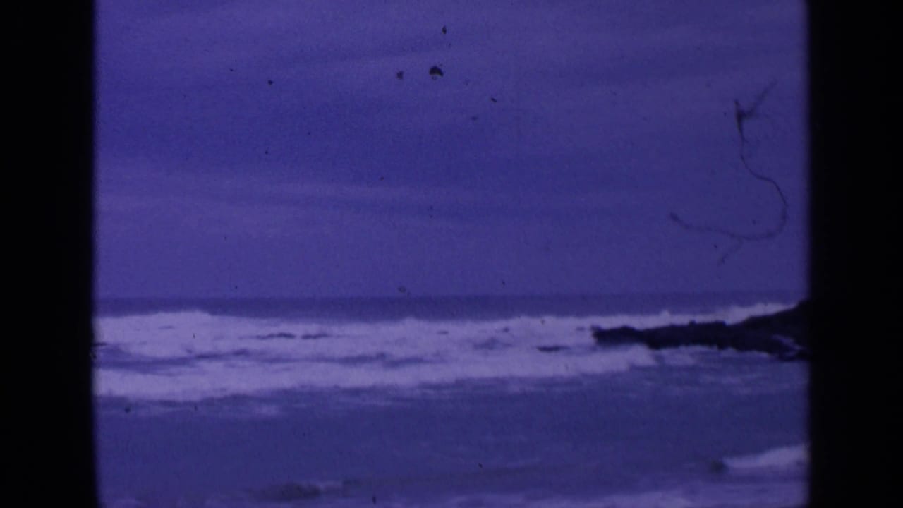 Choppy ocean surf breaks against dark rocks beneath a purple overcast sky.