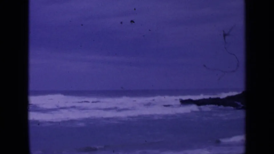 1963 8mm home-movie frame of rough Pacific surf near San Francisco, with dark rocks and a purple color cast.
