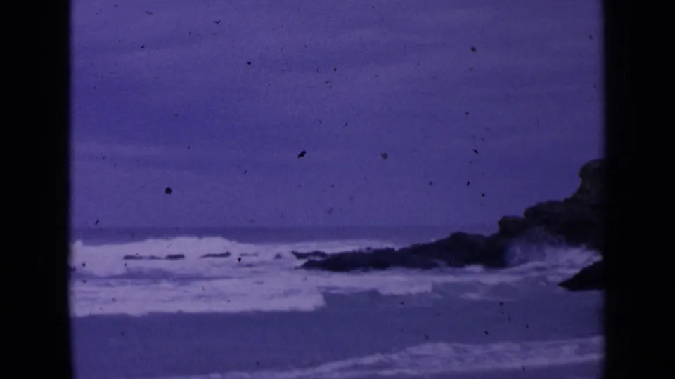 1963 San Francisco coastal home-movie frame with rough waves, dark rocks, grain, dust, and a purple cast.