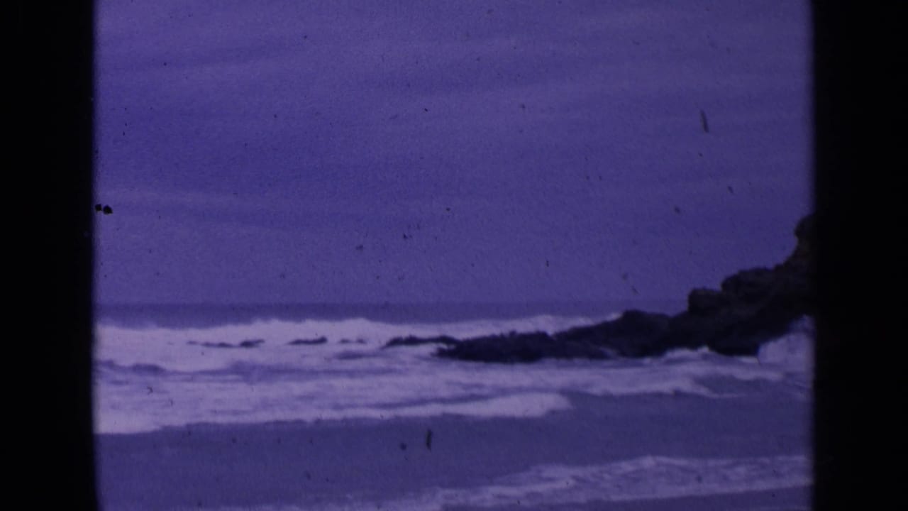 1963 San Francisco 8mm frame showing purple-shifted waves approaching a rocky shoreline beneath an overcast sky.