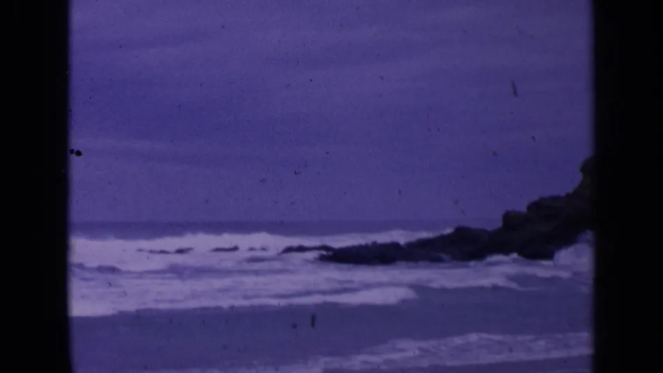 1963 San Francisco 8mm frame showing purple-shifted waves approaching a rocky shoreline beneath an overcast sky.