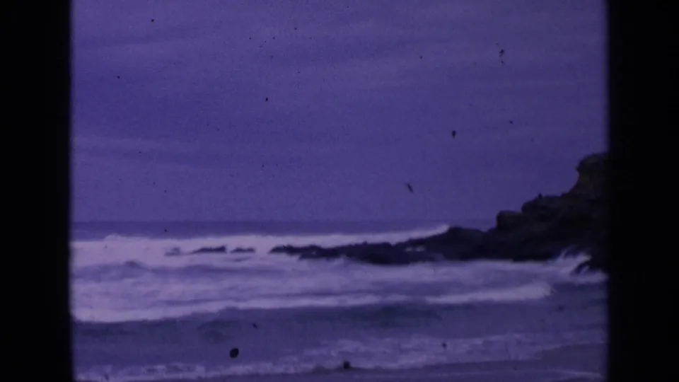 1963 San Francisco coastal frame with waves breaking near dark rocks, soft focus, and visible 8mm dust marks.