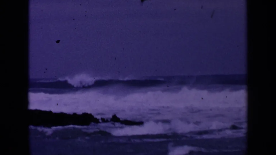 Purple-toned 1963 San Francisco 8mm frame showing low surf, a dark foreground rock line, and a cloudy horizon.