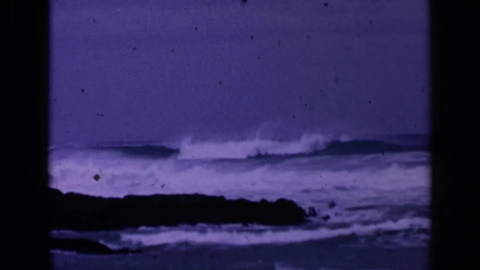 1963 San Francisco shoreline frame with a white wave crest behind dark rocks and visible dust on purple-shifted 8mm film.