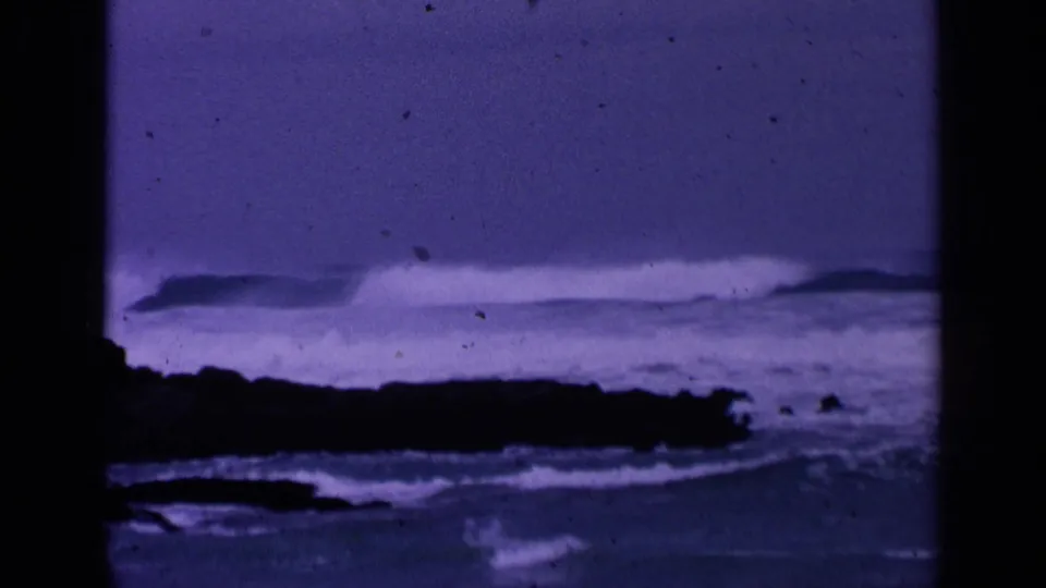 1963 8mm home-movie frame from San Francisco showing surf breaking beyond a dark rocky shoreline under a purple-blue color cast.