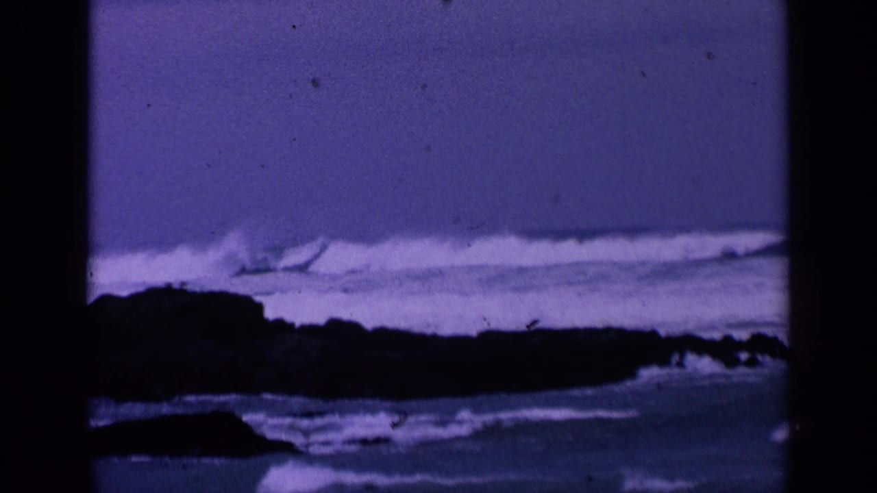 White surf breaking near a dark rocky point with purple film tint.