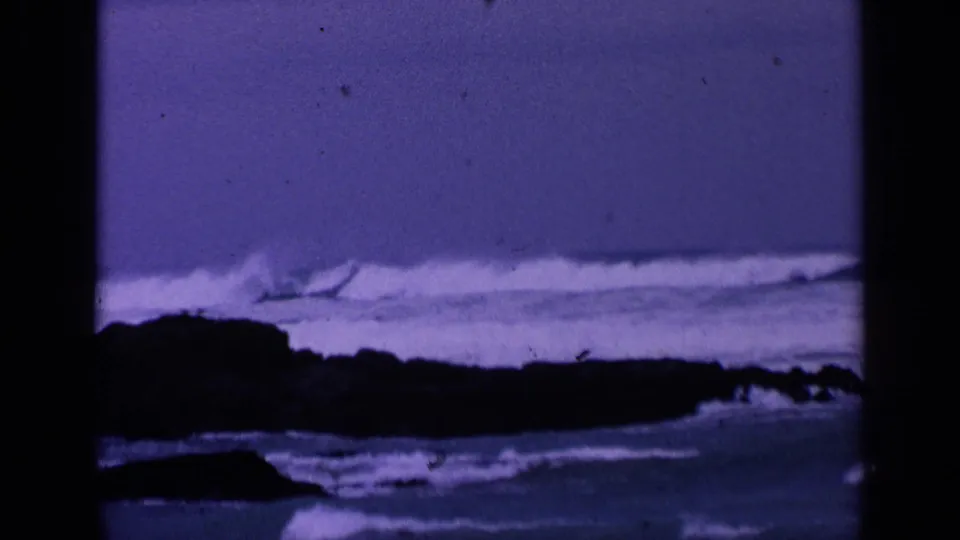 1963 San Francisco coastal frame with white surf, a dark rocky outcrop, and visible 8mm dust and grain.