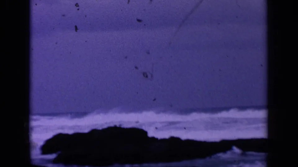 Purple-tinted 8mm frame of a low rocky shoreline with pale surf breaking across the background.