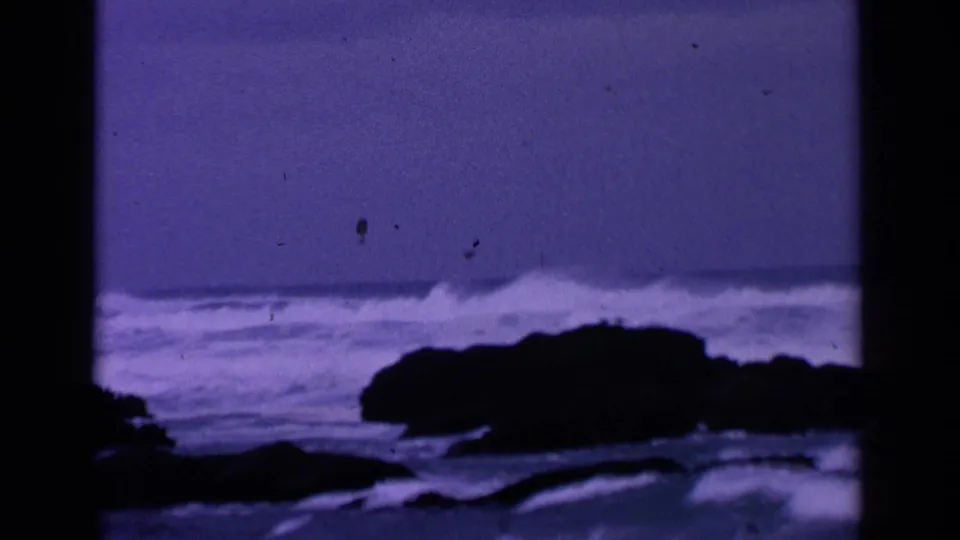 1963 8mm coastal frame showing a dark rock formation in front of foamy Pacific surf with a strong purple-blue cast.