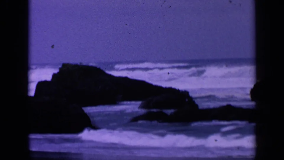 1963 8mm frame of a low rocky California shoreline with pale surf breaking across the foreground.