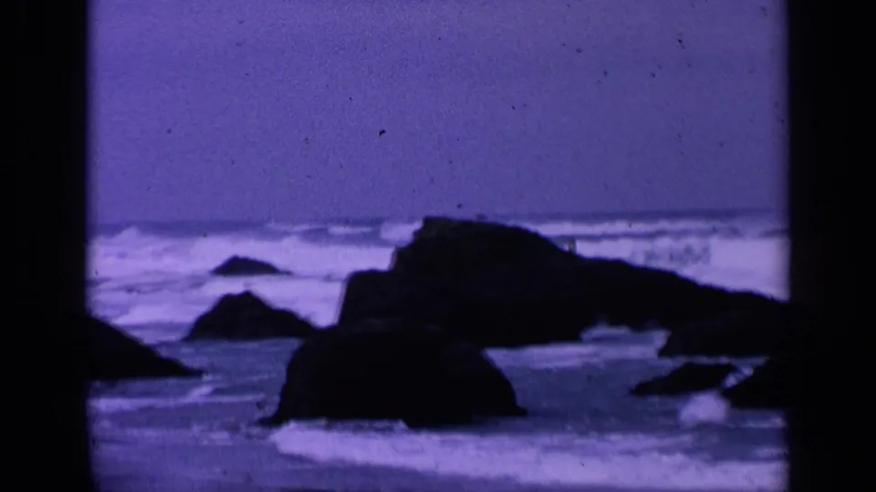 1963 8mm coastal frame near San Francisco with dark rocks, choppy surf, and a hazy purple-blue sky.