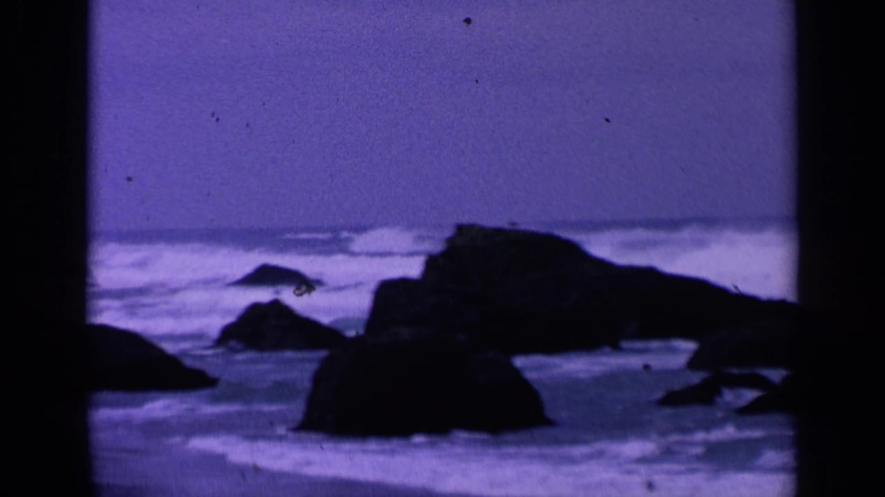 Dark rocks and breaking surf under a purple-blue overcast sky.