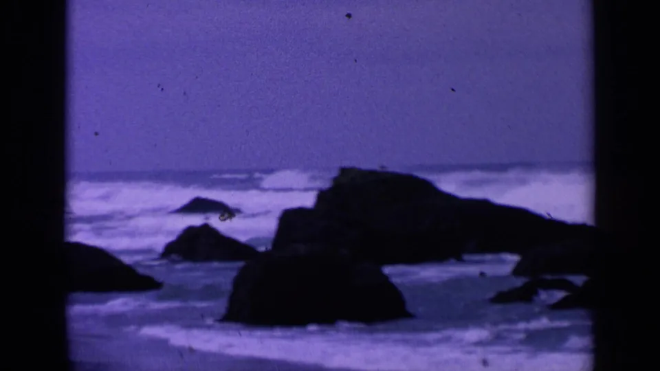 1963 8mm San Francisco coastal view with dark rocks, white surf, and a purple-blue overcast horizon framed by film borders.
