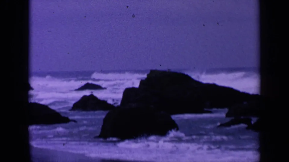 Vintage 8mm view of San Francisco coastal rocks with waves breaking behind them and a deep purple color cast across the frame.