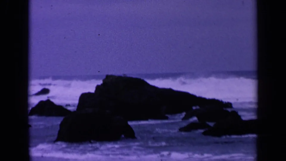 1963 home-movie frame of the San Francisco shoreline, showing dark rocks, foamy surf, and shallow water with visible grain and film edges.