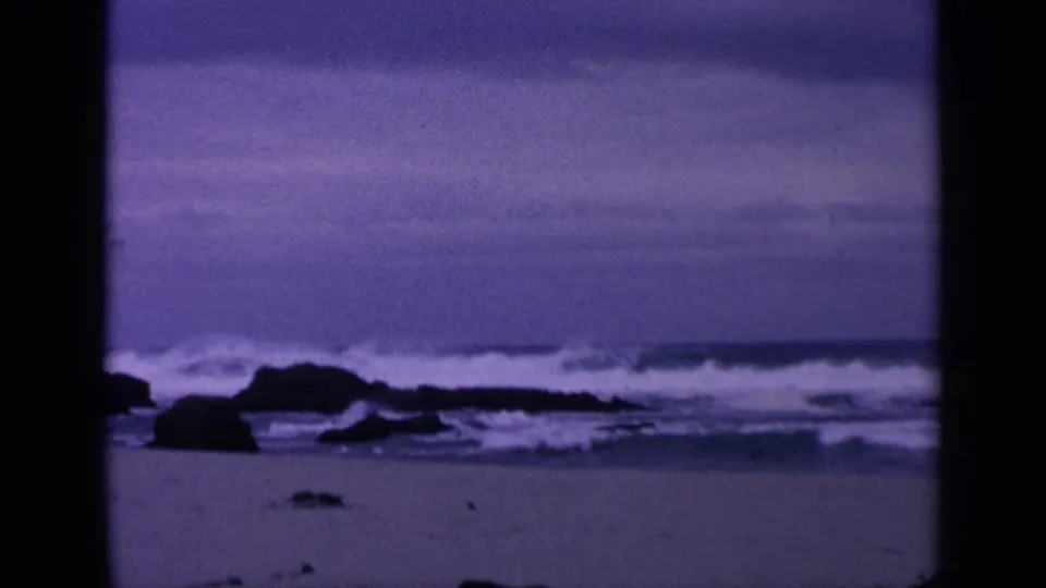 Wide 8mm San Francisco coastal frame with low rocks, breaking surf, wet sand or shallow water, and a cloudy purple-blue sky.